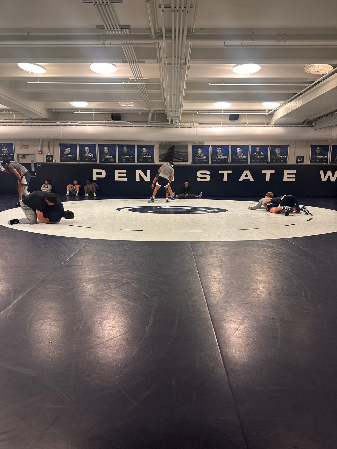 Wrestling room showing several wrestlers on a large Resilite wrestling mat in the center, with PEN STATE written on the wall behind them and a dark blue wrestling mat